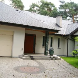 Elegancki dom jednorodzinny z szarą dachówką i beżową elewacją, widok na wejście z kolumnami z zielonego szkła i garaż z bramą segmentową, brukowany podjazd z dekoracyjnym wzorem i rozsypanymi...