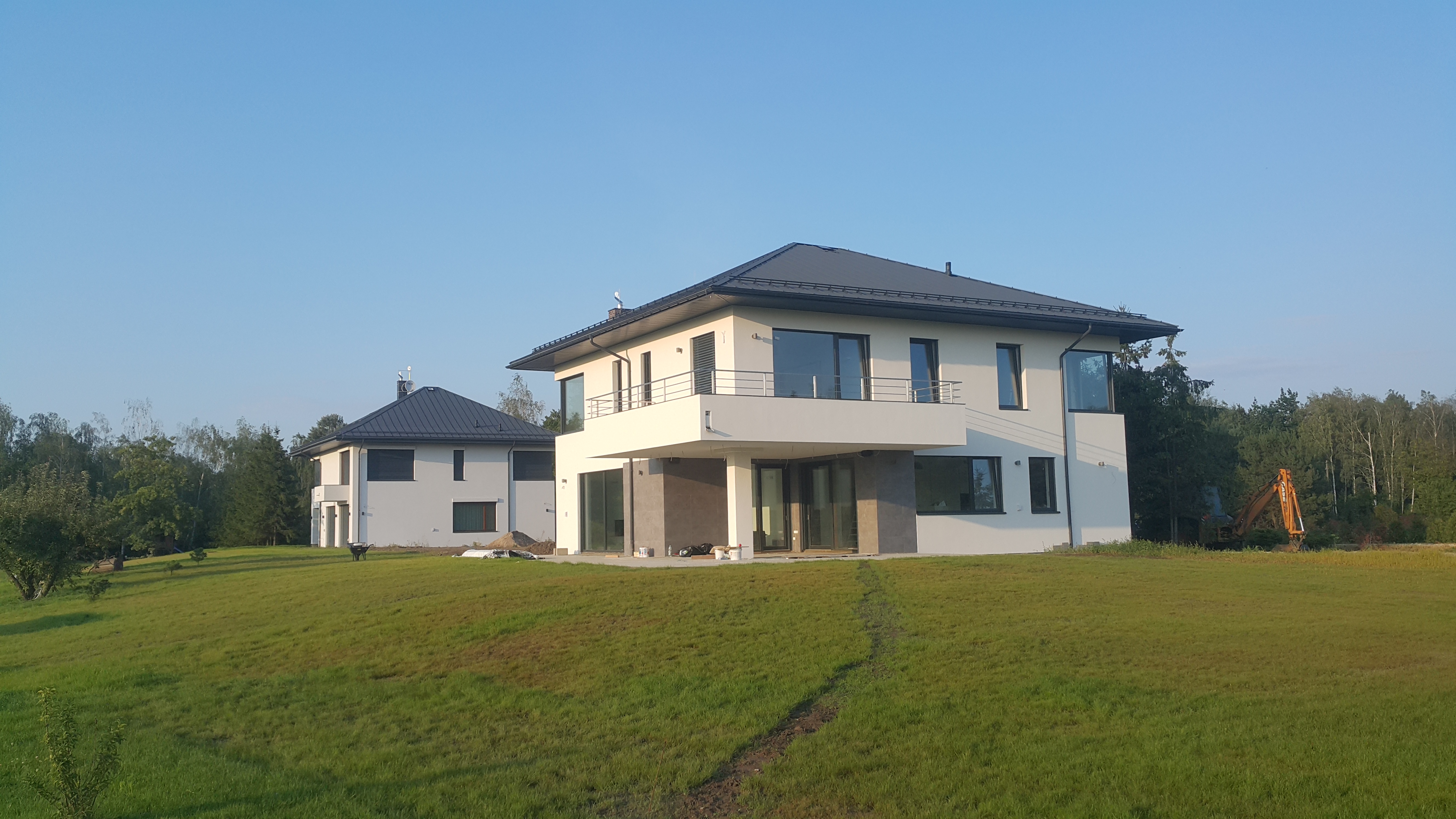 Dwa nowoczesne domy murowane z płaskimi dachami na wzgórzu, z widocznym sprzętem budowlanym w tle, sugerującym trwające prace wykończeniowe.