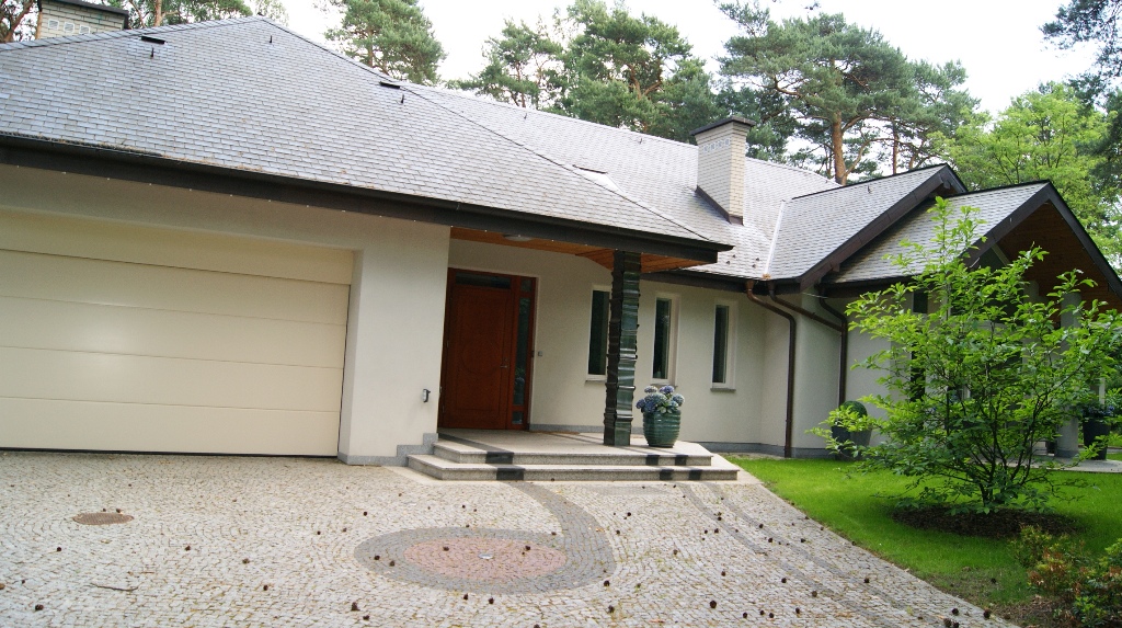 Elegancki dom jednorodzinny z szarą dachówką i beżową elewacją, widok na wejście z kolumnami z zielonego szkła i garaż z bramą segmentową, brukowany podjazd z dekoracyjnym wzorem i rozsypanymi...