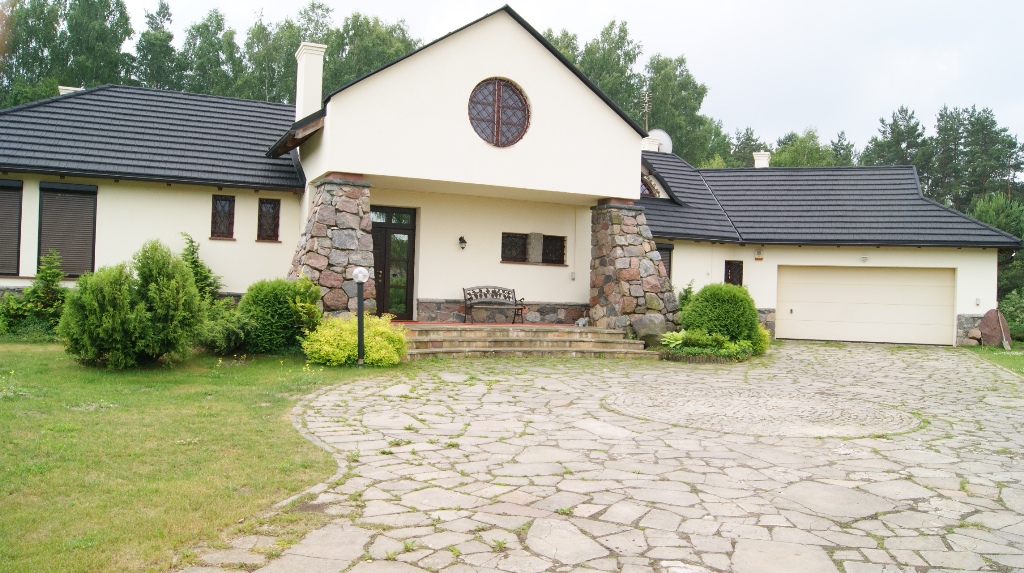 Elewacja domu jednorodzinnego z elementami kamiennymi, okrągłym oknem i czarną dachówką, otoczonego zielenią i brukowanym podjazdem.