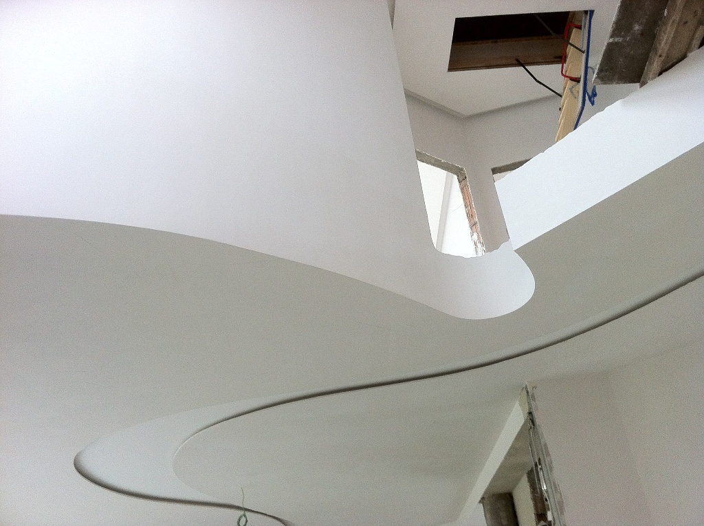 Biały sufit z dekoracyjnymi, falistymi liniami i nietypowymi wycięciami, widoczne elementy konstrukcyjne i instalacje w tle.