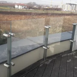 Szklana balustrada balkonowa z metalowymi mocowaniami i drewnianą podłogą, widok na krajobraz miejski w tle.