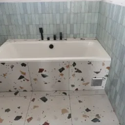 Wykończona łazienka z wanną obudowaną płytkami lastryko, szare płytki na ścianach ułożone pionowo, czarna bateria wannowa.