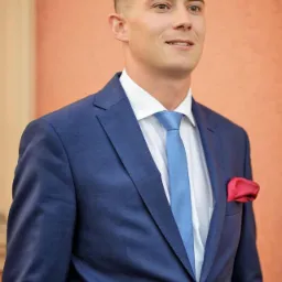 Portret młodego mężczyzny w eleganckim, granatowym garniturze, białej koszuli i błękitnym krawacie, z czerwoną poszetką w kieszonce, spoglądającego w bok.