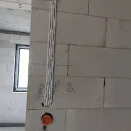 Białe kable elektryczne prowadzone pionowo wzdłuż surowej ściany z betonu komórkowego, z pomarańczową puszką instalacyjną i odręcznymi notatkami ołówkiem na ścianie.