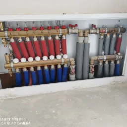 Rozdzielacz ogrzewania podłogowego z rurami w kolorze czerwonym i niebieskim, umieszczony w białej szafce w surowym stanie, z widocznymi numerami na belce rozdzielacza.