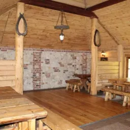 Rustykalna sala z drewnianymi ścianami, sufitami i podłogami, oświetlona wiszącą lampą z koła od wozu, z dekoracjami w postaci podków na słupach i stołami z ławami.