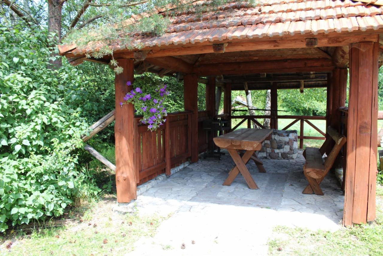 Drewniana altana z rustykalnym stołem i ławką, ozdobiona fioletowymi kwiatami, otoczona zielenią.