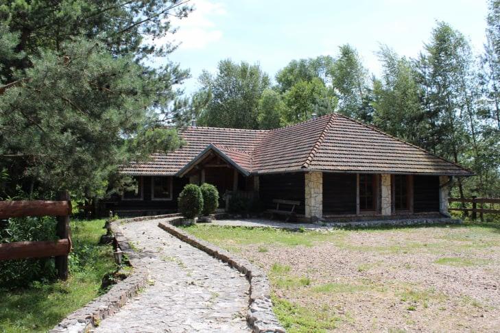 Drewniany budynek z brązowym dachem krytym dachówką, otoczony zielenią, z kamienną ścieżką prowadzącą do wejścia.