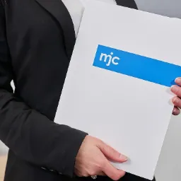 Osoba w czarnym żakiecie trzyma biały folder z niebieskim paskiem i logo 'mjc'.