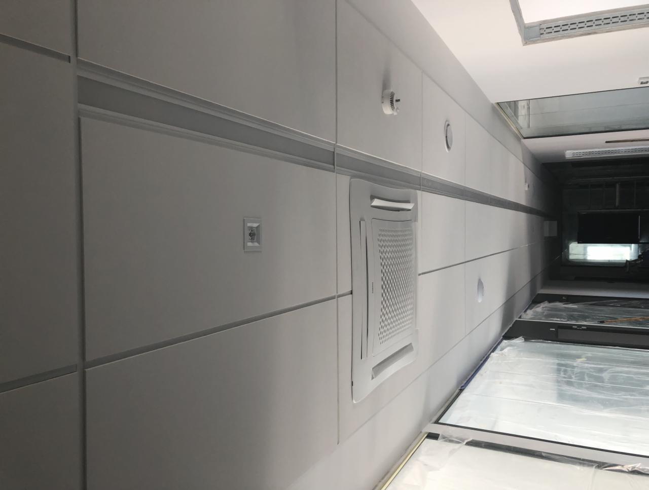 Wykończona ściana z płyt z wbudowanym panelem klimatyzacji, widoczne gniazdko elektryczne i czujnik dymu, całość w odcieniach szarości z delikatnymi listwami dekoracyjnymi.