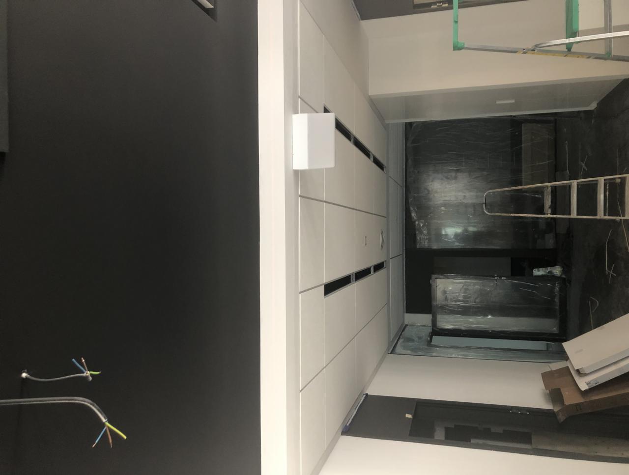 Wnętrze w trakcie prac wykończeniowych: czarna ściana z wystającymi przewodami, białe szafki w zabudowie, folia ochronna na podłodze i elementach wyposażenia, drabina i fragment konstrukcji sufitu...