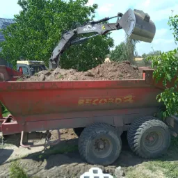 Czerwona przyczepa rolnicza z napisem RECORD załadowana ziemią, z której koparka Mecalac zsypuje urobek; w tle dom z panelami słonecznymi.