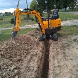 Żółta mini-koparka XN08 wykopuje wąski rów w ziemi, wyznaczony czerwoną linką, z hałdą ziemi obok