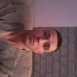 Mężczyzna w szarej bluzie, siedzący na beżowej kanapie, z ceglaną ścianą w tle, obrócony o 90 stopni w prawo.