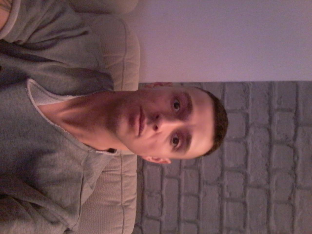 Mężczyzna w szarej bluzie, siedzący na beżowej kanapie, z ceglaną ścianą w tle, obrócony o 90 stopni w prawo.