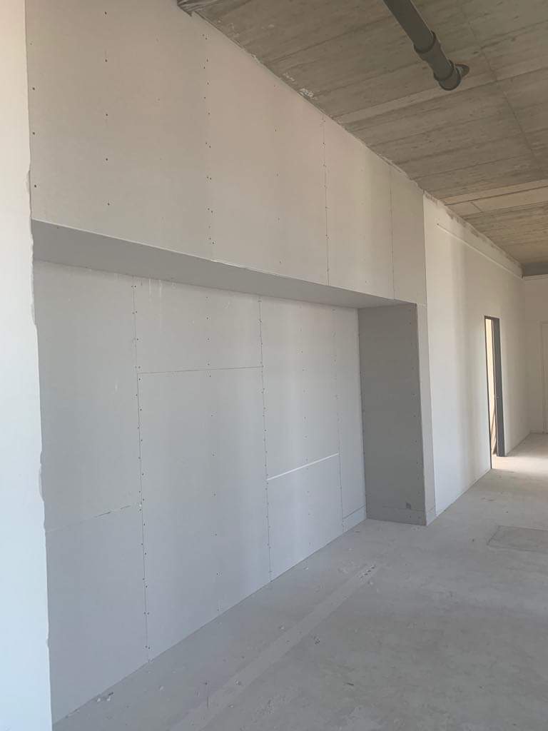 Wnętrze w trakcie prac wykończeniowych: ściana z płyt gipsowo-kartonowych z widocznym wnęką, surowy betonowy sufit z szarą rurą instalacyjną, szara posadzka, widoczne łączenia płyt.