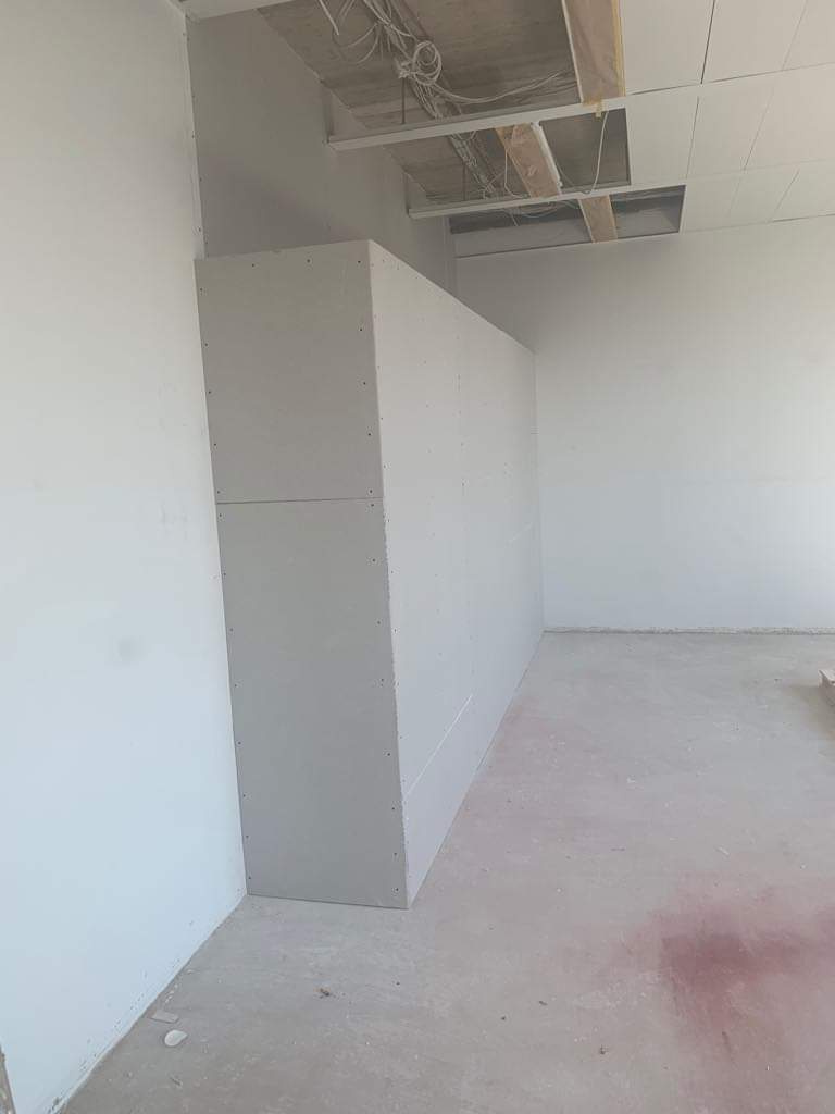 Wnętrze w trakcie prac wykończeniowych: ściana z płyt karton-gips, widoczne łączenia i wkręty, sufit z odsłoniętą konstrukcją i przewodami.