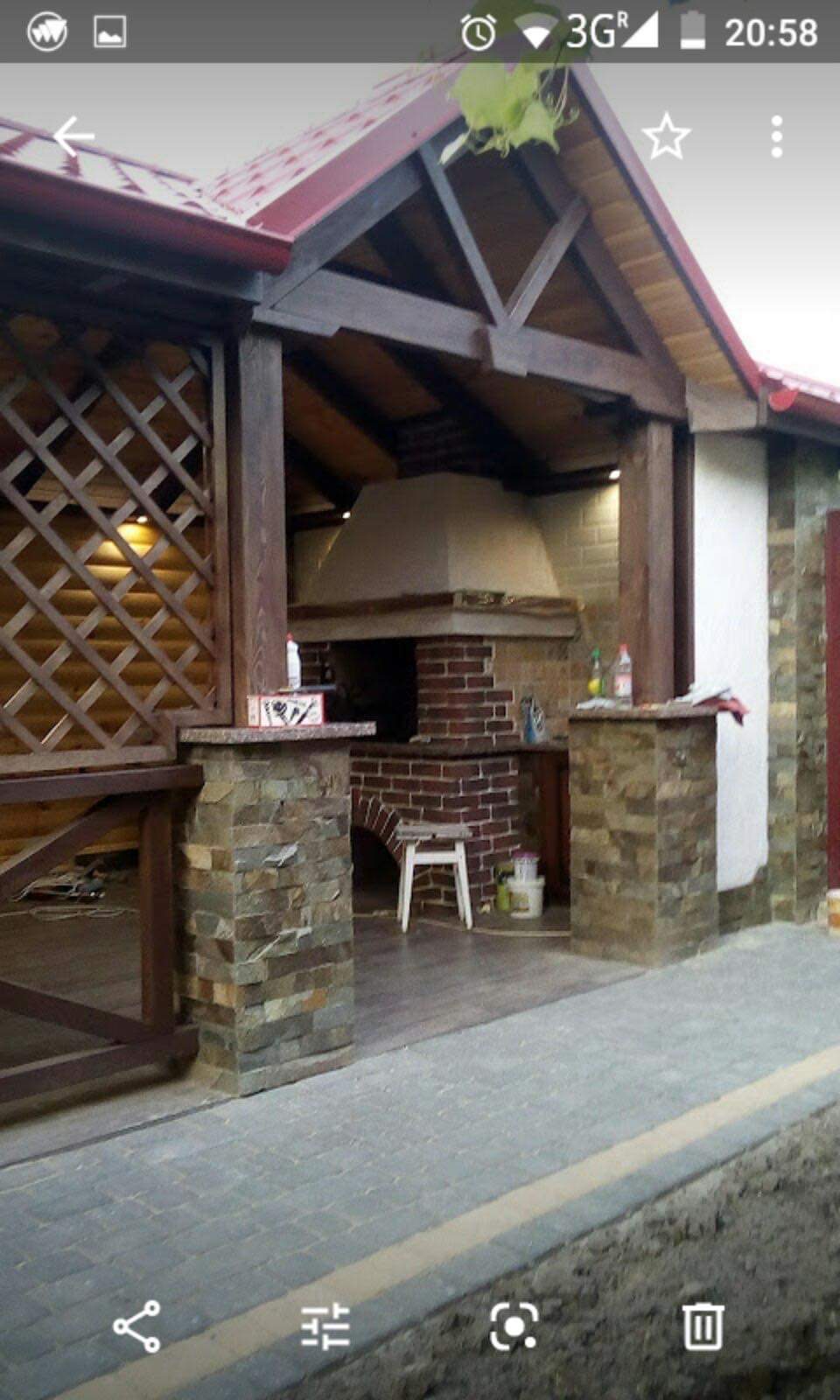 Zadaszona, murowana kuchnia letnia z ceglanym grillem i kamiennymi filarami, widoczna z zewnątrz, z drewnianymi elementami konstrukcyjnymi dachu i kratą na balustradzie.