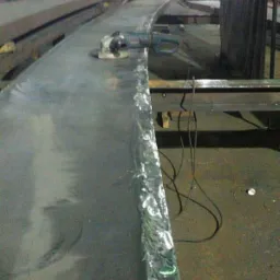 Metalowa płyta z zabezpieczonym brzegiem i szlifierką kątową na powierzchni, w tle hala produkcyjna z widocznymi pracownikami.