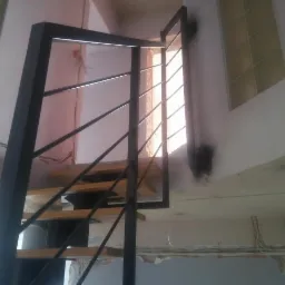 Schody drewniane z czarną metalową balustradą w surowym wnętrzu, widok od dołu, z oknem z luksferów w tle.