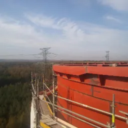 Czerwona, metalowa konstrukcja zbiornika w Sosnowcu, widoczna ze rusztowania na tle lasu, komina elektrowni i linii wysokiego napięcia.