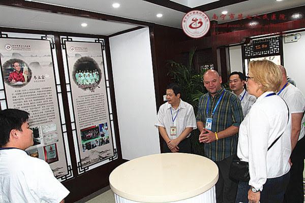 Grupa osób, w tym obcokrajowcy, ogląda tablice informacyjne z tekstem w języku chińskim i zdjęciami w pomieszczeniu przypominającym biuro tłumaczeń lub instytucję kulturalną.