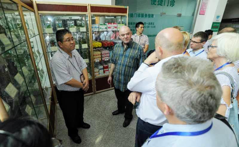 Grupa osób, w tym mężczyzna w niebiesko-zielonej koszuli w kratę, słucha osoby w okularach stojącej obok gablot z eksponatami; w tle widoczny napis w języku chińskim.