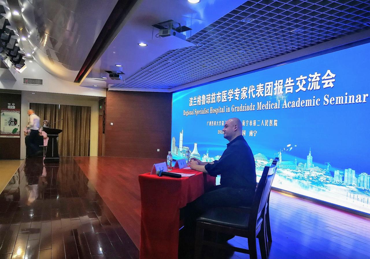 Mężczyzna siedzący za stołem na tle ekranu z informacjami o seminarium medycznym, widoczna woda i mikrofon.