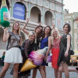 Grupa sześciu uśmiechniętych kobiet pozuje z balonami w kształcie butelki szampana i lodowego rożka na tle zabytkowego budynku z arkadami w Poznaniu.