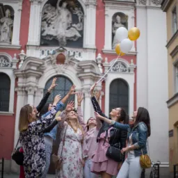 Grupa kobiet w różnym wieku i strojach, stojąca na tle barokowego kościoła, wyciąga ręce ku górze w stronę białych i złotych balonów.