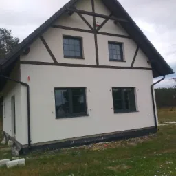 Dwukondygnacyjny dom z jasną elewacją, ciemnymi ramami okiennymi i elementami imitującymi konstrukcję szachulcową na szczycie, widoczne rynny i fragment terenu z resztkami materiałów budowlanych.