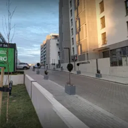 Nowoczesne budynki mieszkalne z biurem sprzedaży w kontenerze, ul. Rudereńskiego 51e, nasadzenia drzew w donicach wzdłuż drogi dojazdowej.