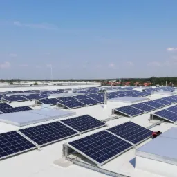 Widok z góry na dużą instalację paneli słonecznych na dachu budynku przemysłowego w słoneczny dzień. Widoczny fragment pracownika w czerwonej koszulce.