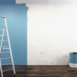 Ściana w połowie pomalowana na niebiesko, z aluminiową drabiną, puszką farby i wałkiem na drewnianej podłodze.