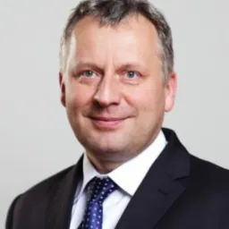 Portret mężczyzny w garniturze z niebieskim krawatem w kropki, uśmiechającego się do kamery na jasnym tle. Fotografia biznesowa z subtelnym oświetleniem.