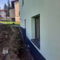 Fundament budynku zabezpieczony czarną folią hydroizolacyjną, drenaż z kamieni, wykop w ziemi. Łopata oparta o ziemię. Widok z boku budynku.