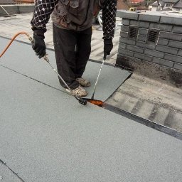 Cinek Marcin Barzowski - Dekarz łączy papę termozgrzewalną na dachu za pomocą palnika, widoczny komin z szarej cegły i fragmenty zabudowy mieszkalnej w tle.