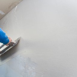 Cinek Marcin Barzowski - Nakładanie gładzi gipsowej na ścianę za pomocą szpachelki przez osobę w niebieskiej bluzie, widoczne nierówności i surowy stan wykończenia.