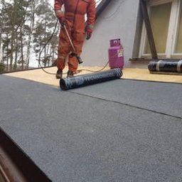 Cinek Marcin Barzowski - Dekarz w pomarańczowym kombinezonie podczas prac na dachu pokrytym czarną papą termozgrzewalną, obok butla z gazem, widoczne drzewa w tle.