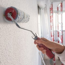 Cinek Marcin Barzowski - Malowanie ściany wałkiem z chropowatą strukturą, widoczna drabina w tle, ręka w ubrudzonej farbą bluzie trzymająca wałek z czerwonym uchwytem.