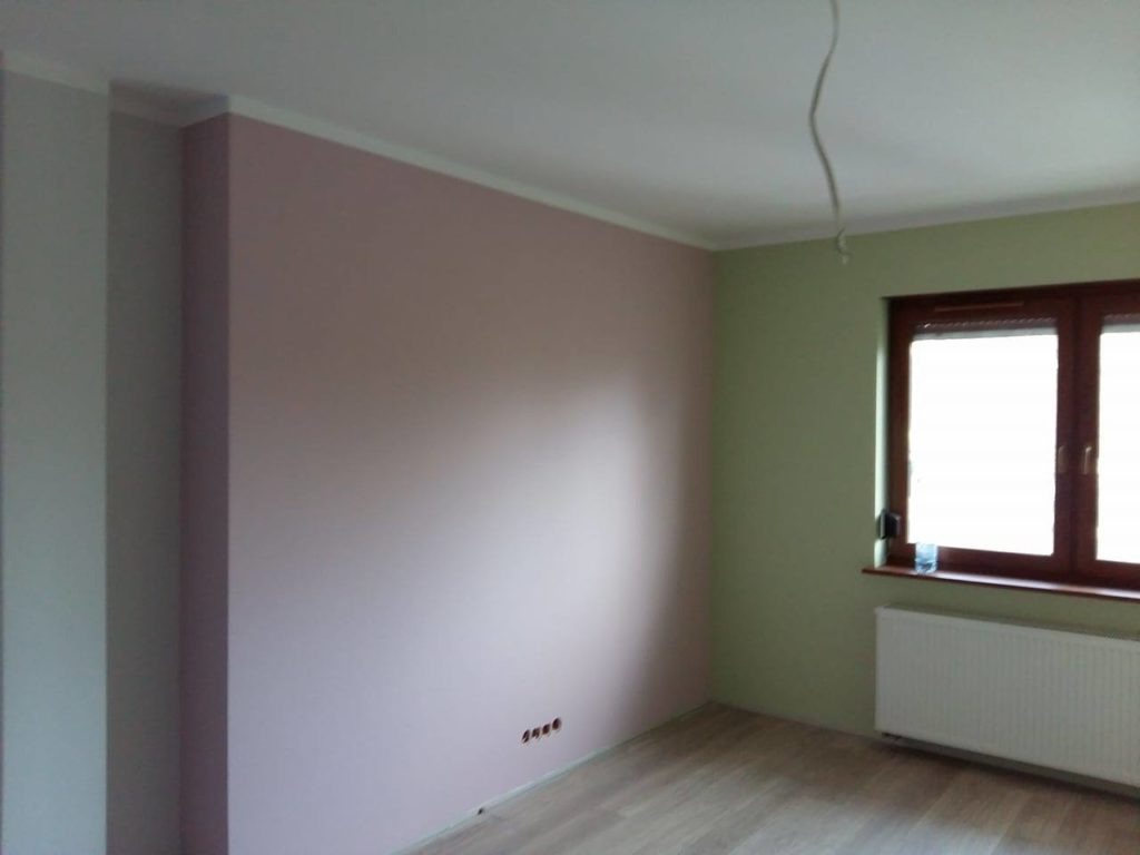Wnętrze pokoju po remoncie: ściany pomalowane na różowo i jasnozielono, drewniana rama okienna, grzejnik pod oknem, drewniana podłoga, wystający kabel ze ściany.