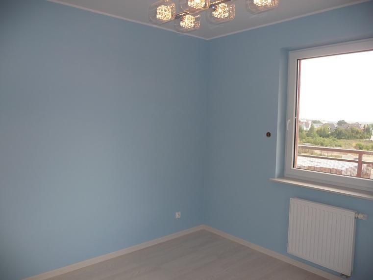 Pomalowane na błękitno wnętrze pokoju z grzejnikiem pod oknem i nowoczesnym oświetleniem sufitowym.