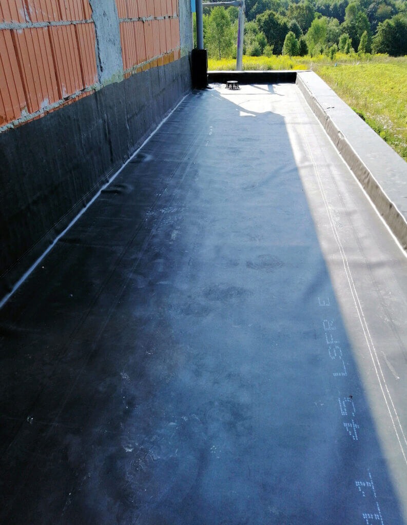 Hydroizolacja płaskiego dachu z papy bitumicznej, widoczne łączenia i fragment ściany budynku w trakcie budowy.