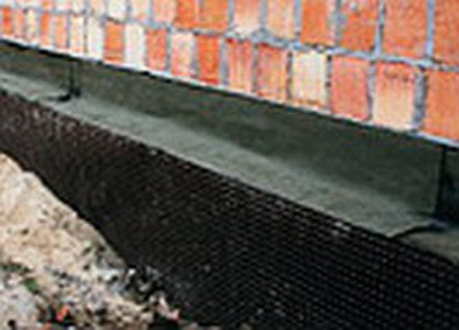 Fundament budynku z izolacją pionową w trakcie prac ziemnych, widoczna siatka drenażowa i ceglana ściana.