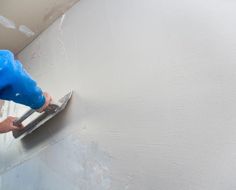 Nakładanie gładzi gipsowej na ścianę za pomocą szpachelki przez osobę w niebieskiej bluzie, widoczne nierówności i surowy stan wykończenia.