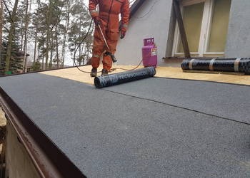 Dekarz w pomarańczowym kombinezonie podczas prac na dachu pokrytym czarną papą termozgrzewalną, obok butla z gazem, widoczne drzewa w tle.