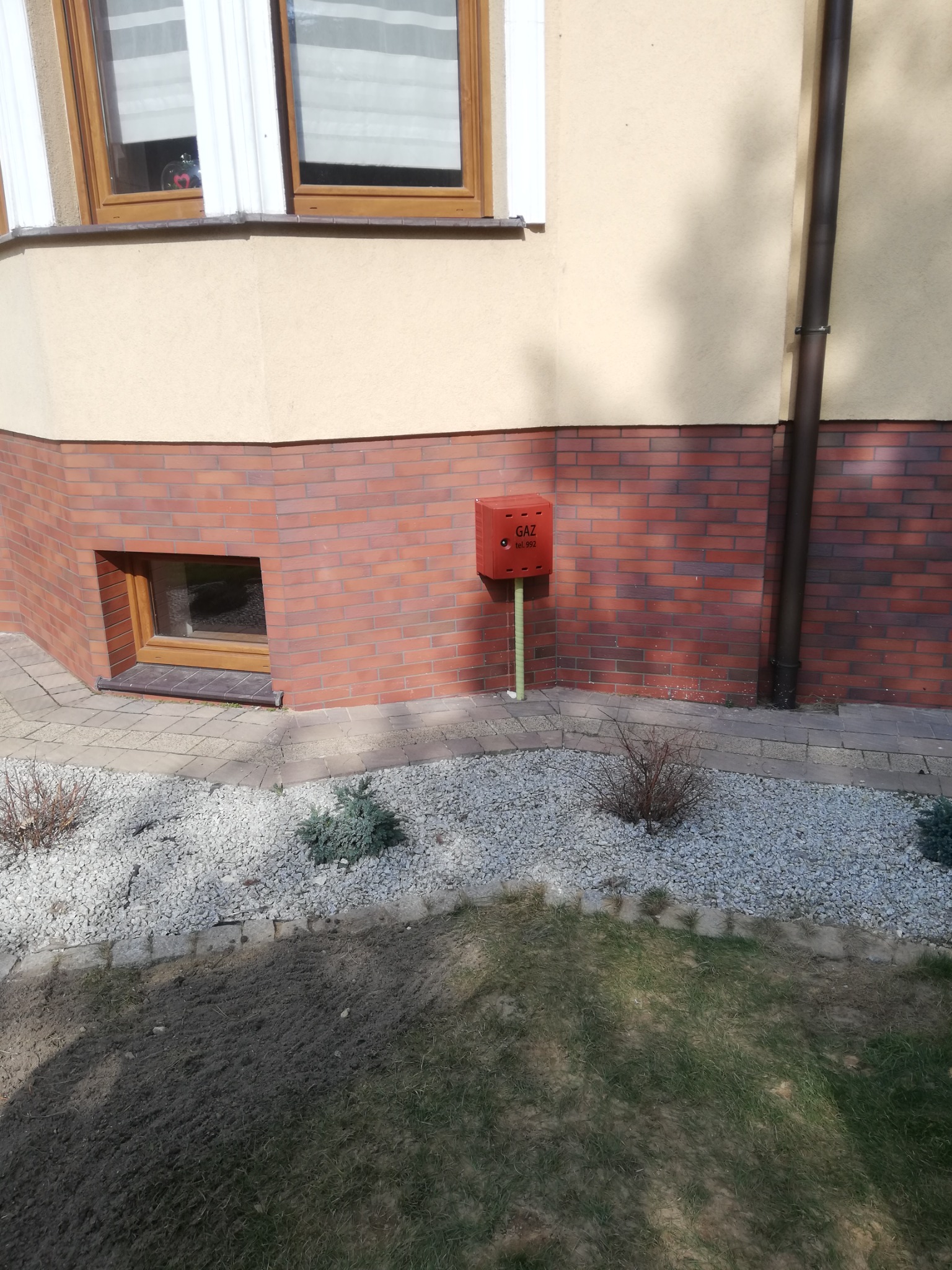 Czerwona skrzynka gazowa z napisem 'GAZ' i numerem telefonu na ceglanej ścianie budynku, widoczna z poziomu trawnika i kamiennego podłoża.