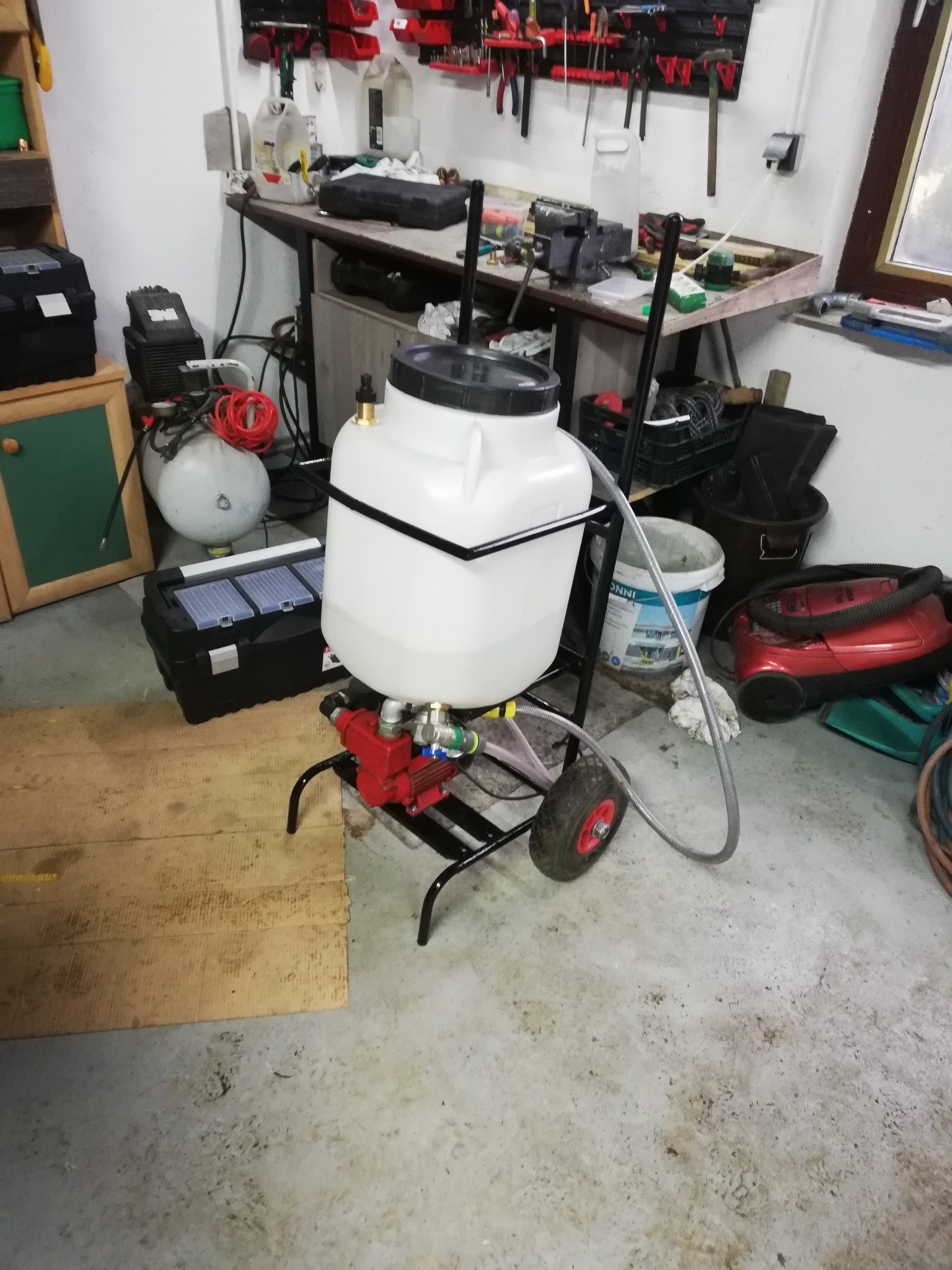 Mobilna stacja robocza z białą zbiornikiem, czerwoną pompą i szarym przewodem na tle warsztatu z narzędziami i akcesoriami.