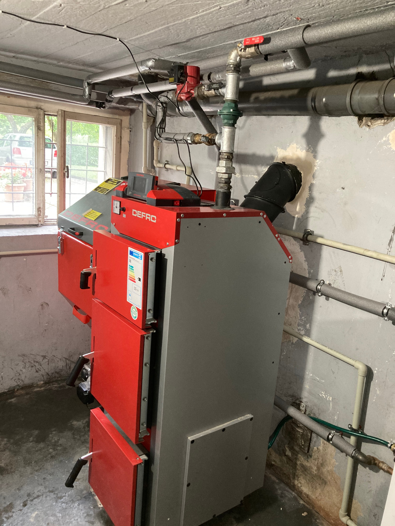Czerwono-szary piec CO marki Defro w piwnicy, widoczne rury instalacji grzewczej i okno z widokiem na zewnątrz.